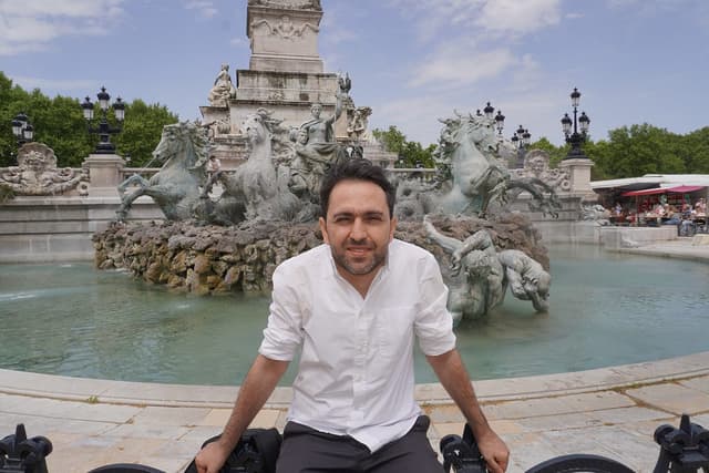 Gevorg Minasyan sits on the edge of classical sculpture fountain on a partly cloudy day.