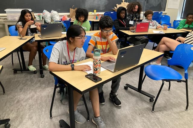 Seven students using their laptops in a classroom. One person is helping two of the students.