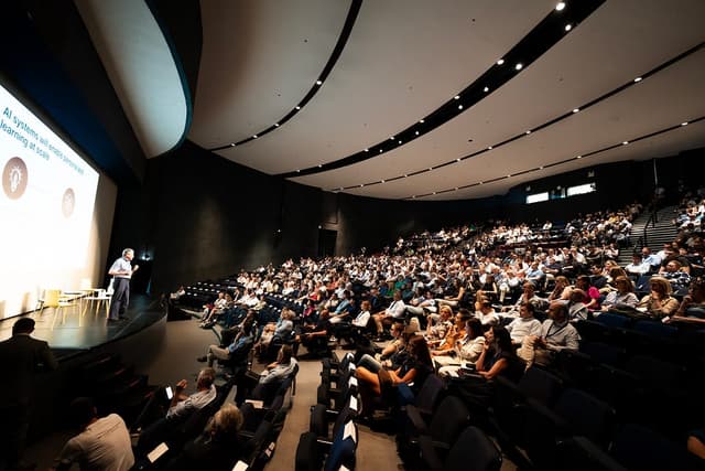 Candid photo of a conference setting, where one speaker is standing onstage with a presentation displayed behind him on a large screen, in front of an audience of several hundred people.