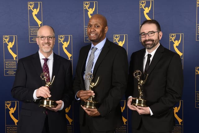 Three people smile at the camera holding Emmy Awards.