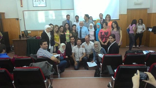 Twenty-four people stand in front of a classroom with a whiteboard in the background.