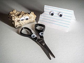 Photo of a beige-colored, bumpy rock, a piece of lined paper, and a pair of scissors, all with googly eyes attached to them.