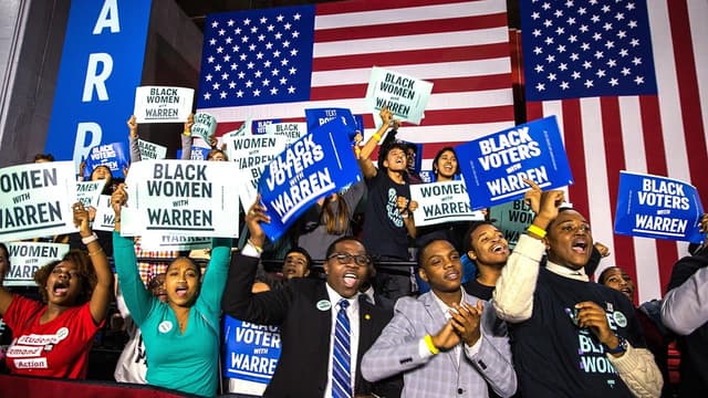 A group of people gather in front of a large U.S. flag, holding signs reading “Black Women with Warren” and “Black Voters with Warren.”