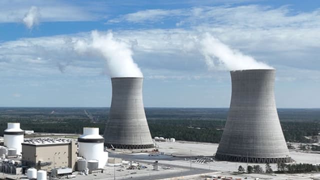 Two large concrete cooling towers at a power plant release white steam into a blue sky, with industrial buildings and storage tanks in the foreground.
