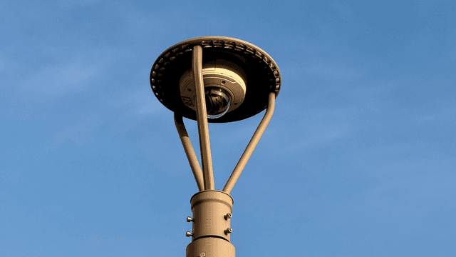 A surveillance camera atop a metal pole, against a blank blue sky.