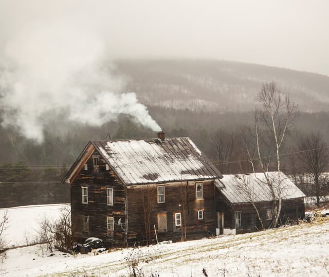 A dark wood clad house with snow visible on a standing-seam metal roof. Smoke is coming from the chimney. Snow-covered fields and winter forest are in the background.