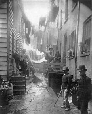 Men in bowler hats stand in the alleyway of a tenement house. Laundry hangs on lines above them.