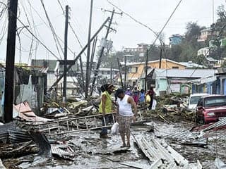 A photo of two men standing in debris after Hurricane Maria hit Puerto Rico. They are surrounded by damaged houses and fallen telephone poles and wires.