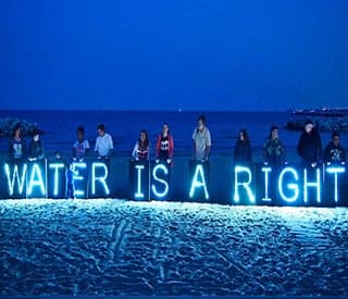 Protesters stand on a beach at nightfall with a neon blue sign that reads “Water is a right”.