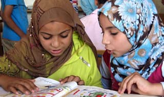 Two girls in headscarves read together from a book.