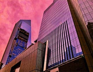 Two glass buildings rise into the sky. They glow pink as they reflect the sunset.