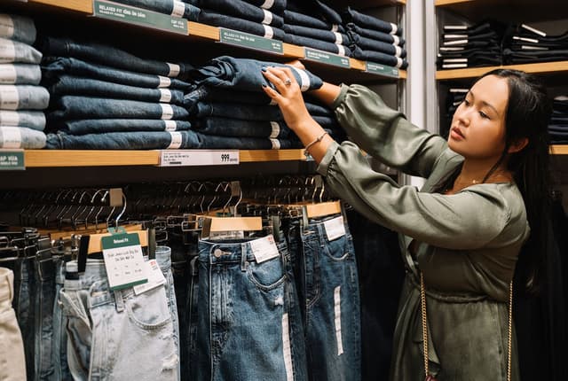 A woman in an olive green top is browsing a stack of dark wash denim jeans on a wooden shelf.