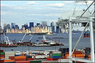 A photograph with the skyline of New York in the background and dozens of colorful shipping containers in the foreground. Ships loading and offloading float in the water.