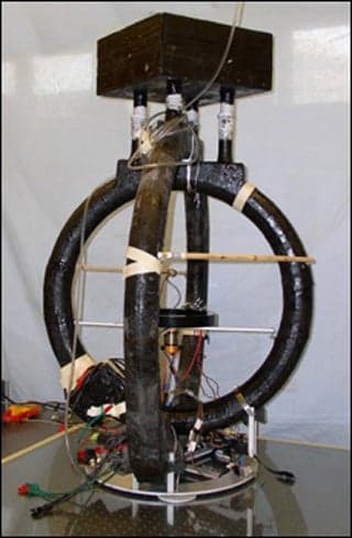 A photo of an electromagnetic device. It looks like a spokeless unicycle with another ring crossecting it and wires connected to it.