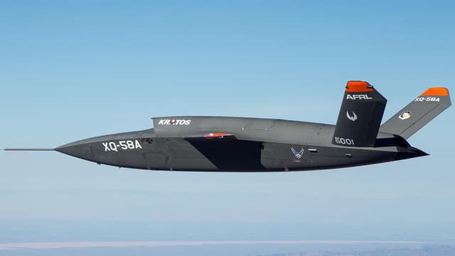 A sleek gray plane with a pointed nose and orange-tipped wings flies against a clear, blue sky.