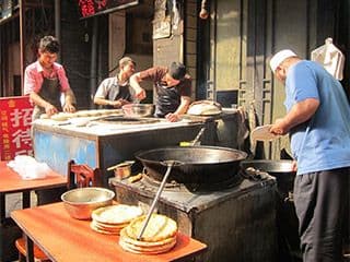 One man stands near a huge pot while three other men prepare food for cooking.