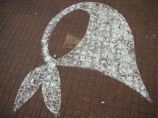 An image of a white shawl tied at the neck is superimposed on a red brick background.
