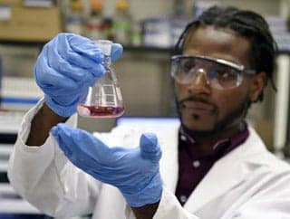 A young male, wearing goggles and gloves, observes purple liquid in a beaker.