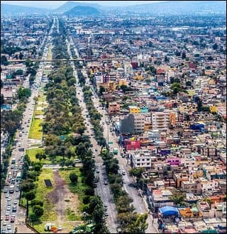 An aerial photograph showing buildings crowded together on the right side with mountains in the background. A long thin strip of green runs parallel to the roads.
