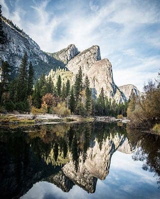 A photograph of a rock formation known as "Three Brothers" at Yosemite National Park. The rock formation is surrounded by trees and reflected in a body of water in the foreground.