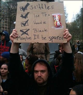 A young white man wearing a hoodie holds a sign over his head with the words "Skittles," "Ice tea," and "Hoodie" listed next to boxes with check marks. Beneath them, next to a box with an x marked in it , are the words "Black. Maybe I'll be spared."
