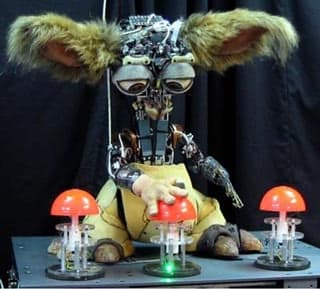 A photograph of the robot Leo. Leo stands before three plunger style buttons with its hand on the center button.