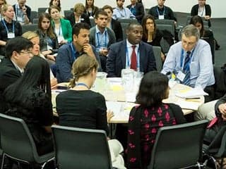 A group of younger and older adults of different races and genders, sitting around a circular table, with others watching from chairs around the periphery.