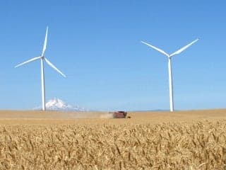 A field of wheat being harvested by a combine, with three wind turbines and a snowcapped mountain in the background.