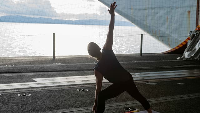 Photo of a person doing a yoga pose while standing on a mat in the hangar bay of an aircraft carrier ship.