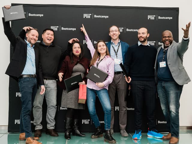 A group of people stand in front of a black background with the words MIT Bootcamps on it.  They each hold a diploma portfolio.