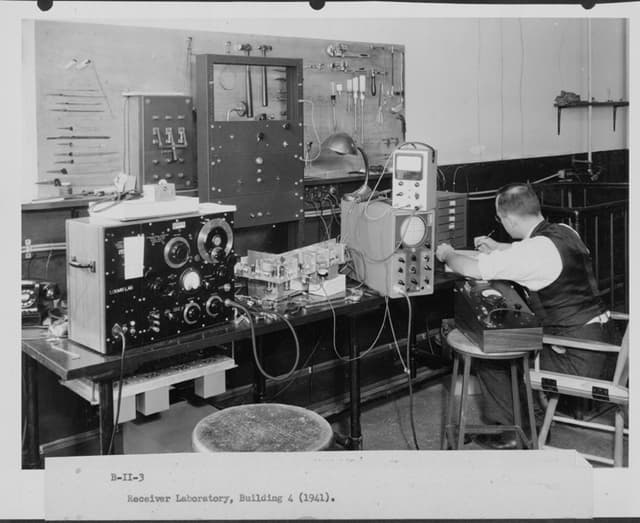 A man writes at a desk which has various large pieces of radio equipment.