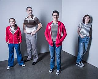 A man with a stern expression stands amongst three females: a preteen, a teenager, and an adult. Each female wears jeans, sneakers, and a striped shirt.