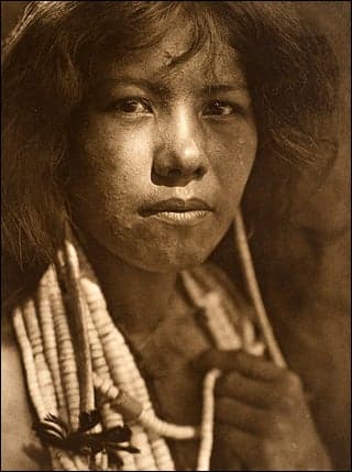 A sepia-toned photograph of a Native American young woman. She has beaded necklaces wrapped around her neck and is looking directly into the camera.